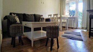 Een woonkamer met een bank, een witte salontafel, twee rieten krukjes en een gestreept vloerkleed.