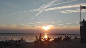 Een strandscène met mensen die ontspannen in stoelen en ligbedden, een vlag en een ondergaande zon boven het water.