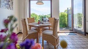 Zona pranzo con sedie in vimini e un tavolo apparecchiato per due, ampie finestre con vista su vegetazione e una porta scorrevole in vetro che dà all'esterno.