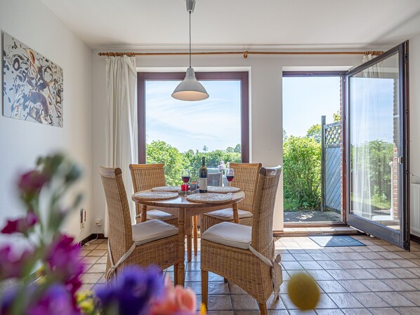 A dining area with wicker chairs and a table set for two, large windows with a view of greenery, and a sliding glass door leading outside.