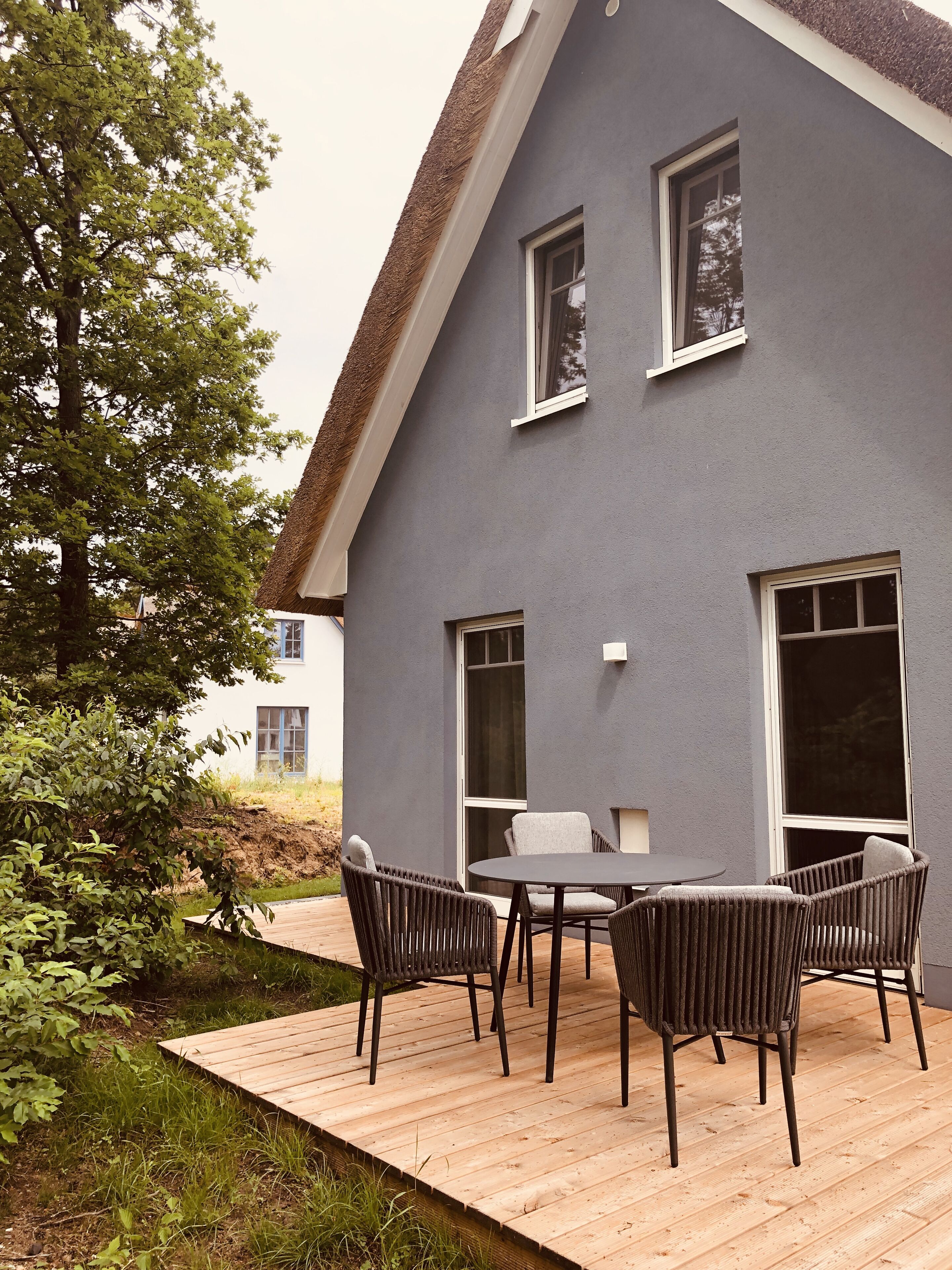 Une maison moderne avec une façade grise, une terrasse en bois et un coin repas comprenant quatre chaises et une table.
