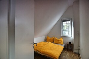 A small, triangular bedroom with a sloped ceiling, a single bed with a yellow blanket, a window with a view of trees, and a small bedside table with a lamp.