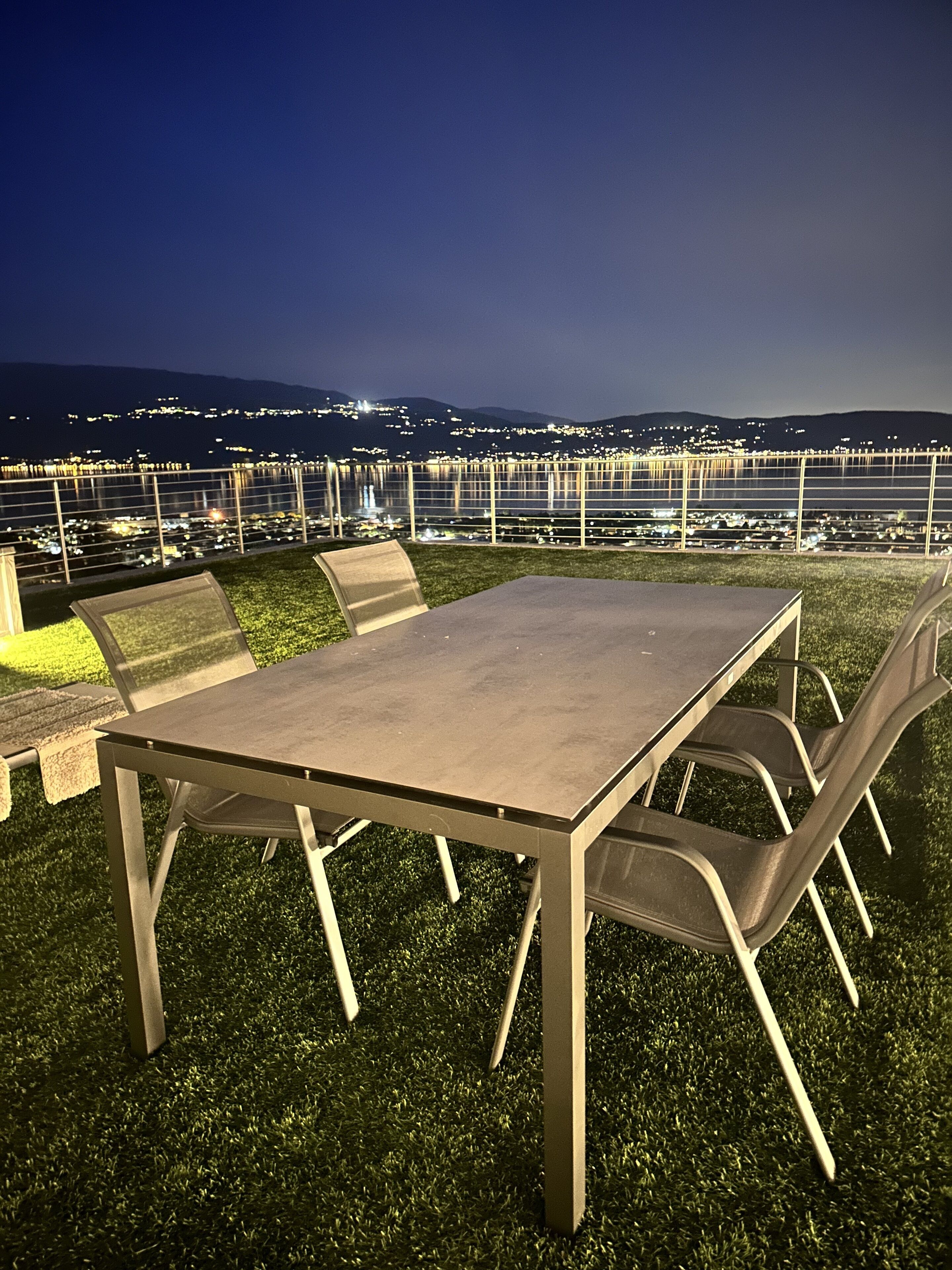An outdoor dining area with a table and chairs, a view of a cityscape at night, and a railing in the background.