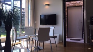 A modern living room with a flat-screen TV, a dining area with chairs, and a large window offering a view of greenery. - Design apartment - 100 sqm green terrace - Pool - Fantastic panoramic lake view (Toscolano-Maderno)