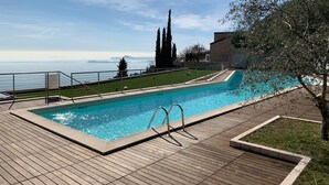 A swimming pool with a wooden deck, surrounded by a fence, and overlooking a body of water with distant land. - Design apartment - 100 sqm green terrace - Pool - Fantastic panoramic lake view (Toscolano-Maderno)