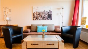 Una sala de estar con un sofá, dos sillones, una mesa de centro y un cuadro de un paisaje urbano en la pared.