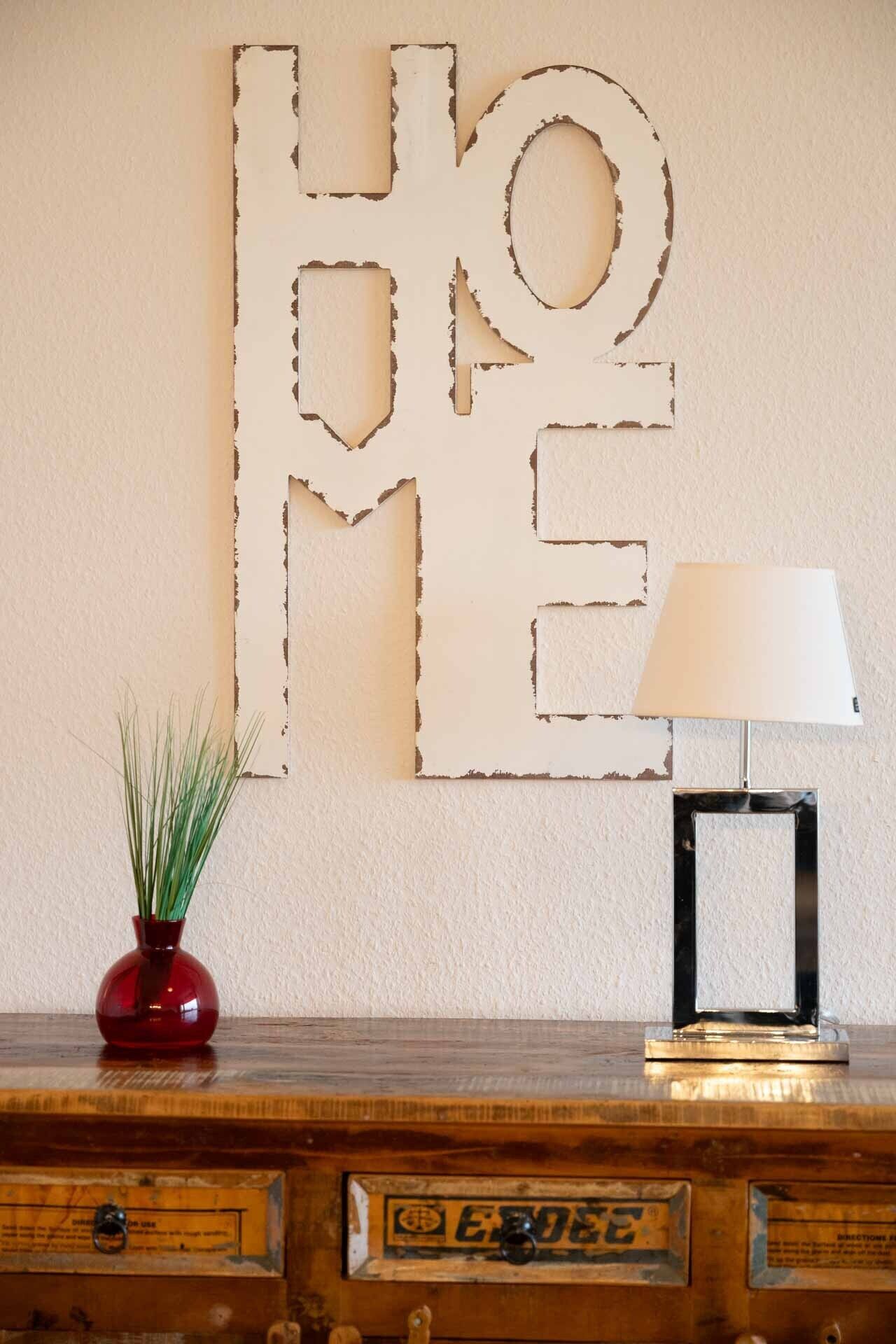 Una pared con una gran decoración abstracta que dice 'HOME', un florero rojo con plantas verdes y una lámpara de mesa sobre un mueble de madera.