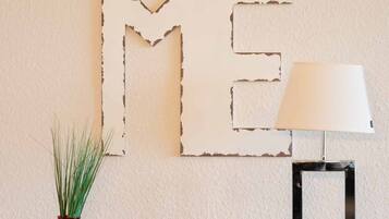 Una pared con una gran decoración abstracta que dice 'HOME', un florero rojo con plantas verdes y una lámpara de mesa sobre un mueble de madera.