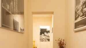 Un pasillo con suelo de madera, una silla amarilla y un jarrón con flores secas. En las paredes hay cuadros enmarcados.