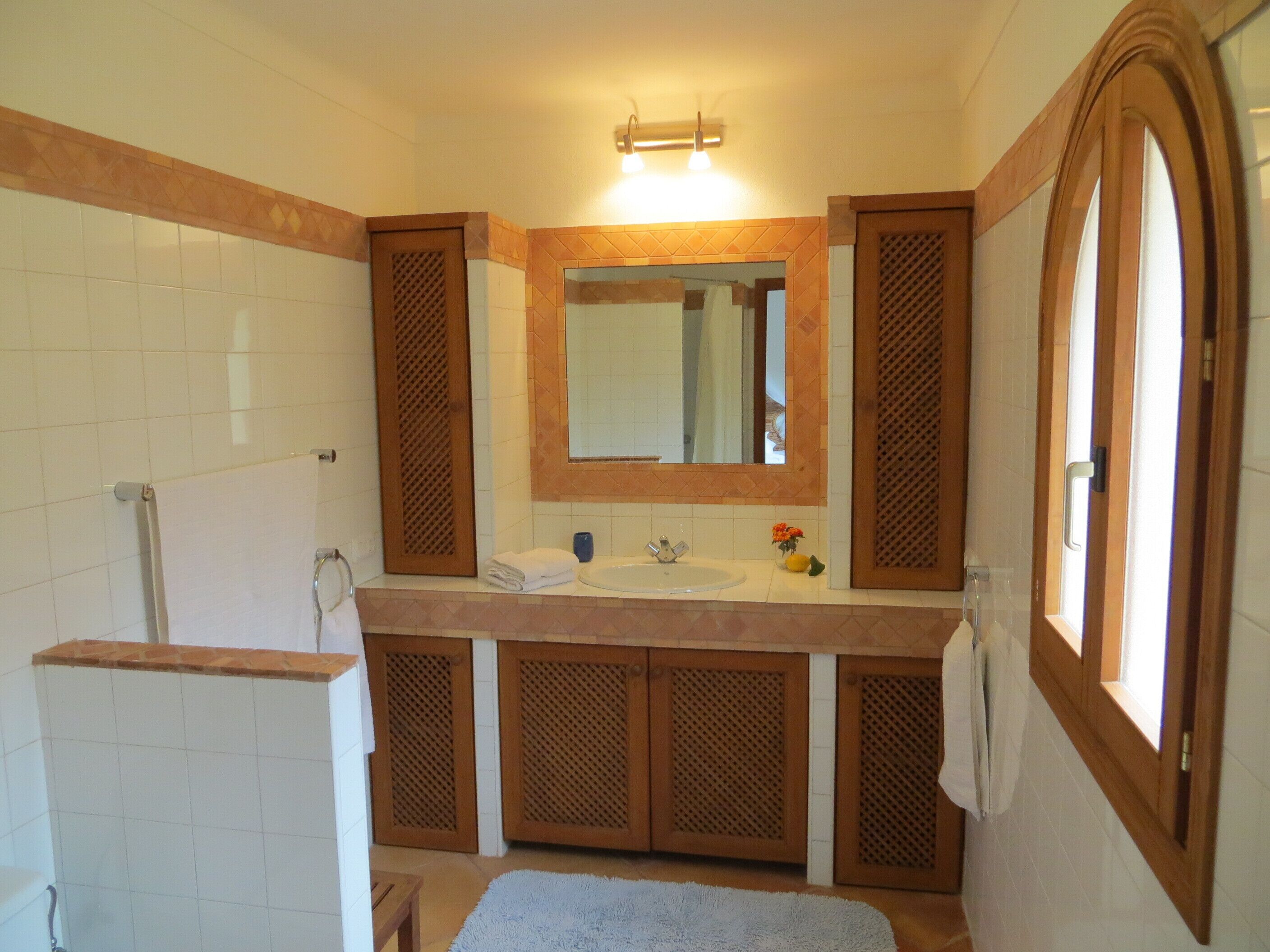 Un baño con pared de azulejos blancos, un mueble de tocador de madera con un lavabo, un espejo sobre el mueble y una ventana a la derecha.
