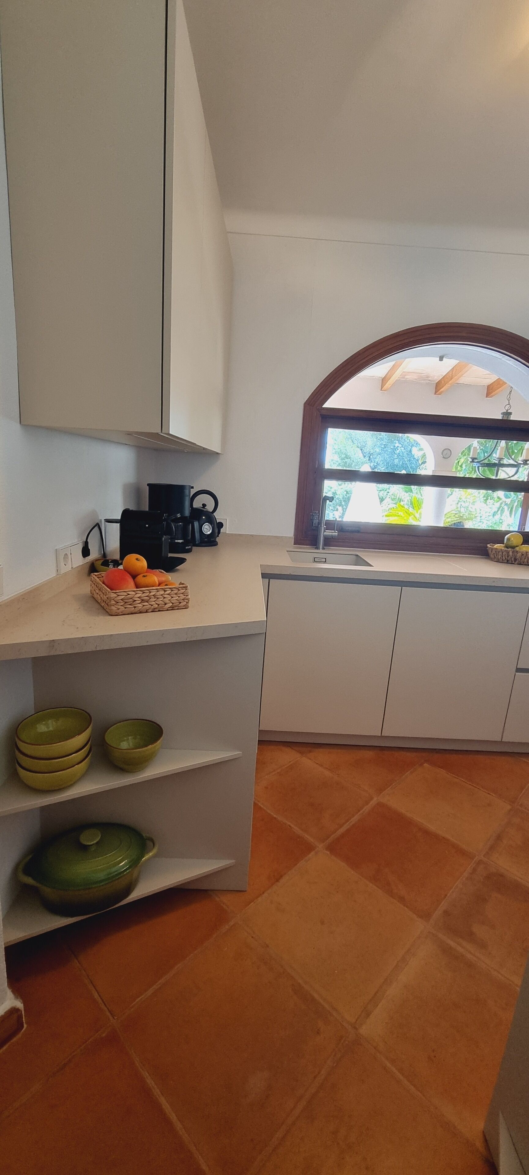 A kitchen with white cabinets, a countertop with a basket of fruit, a coffee maker, and a window with a view of greenery.