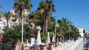 A seaside promenade with palm trees, outdoor seating, and street lamps. - Living room with balcony and sea view, waterfront and south-facing (Vera)