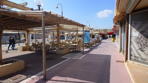 An outdoor café with wooden tables and chairs, a covered seating area, and a street view with shops and a pedestrian walkway. - Beautiful, lovingly furnished holiday home near the beach with sea views (Narbonne)