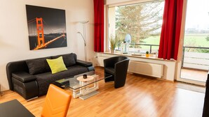 Un salon moderne avec un canapé noir, un fauteuil orange, une table basse en verre et une grande fenêtre ornée de rideaux rouges.