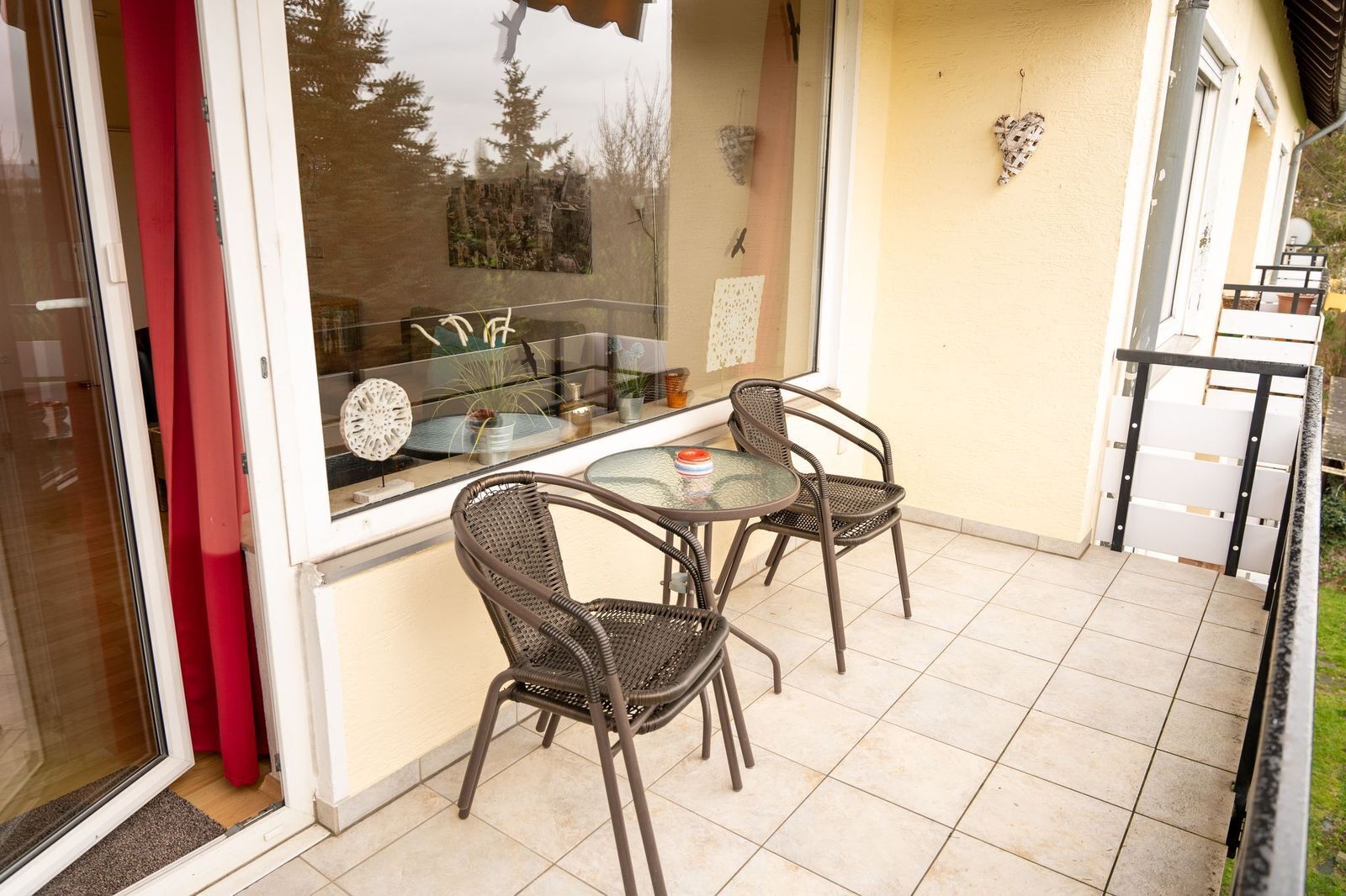 Eine kleine Terrasse mit zwei Korbstühlen und einem Glastisch, eine Glasschiebetür und Blick auf Bäume durch das Fenster.