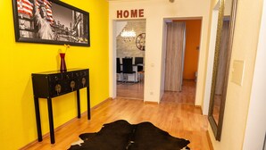 Une pièce avec un plancher en bois, un tapis noir et blanc, une console sur laquelle est posé un vase, un tableau encadré représentant la Statue de la Liberté et un panneau indiquant le mot «MAISON».