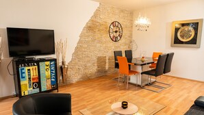 Un salon moderne doté d’un téléviseur à écran plat, d’un mur décoratif en pierre, d’un coin repas avec des chaises orange et d’un lustre.