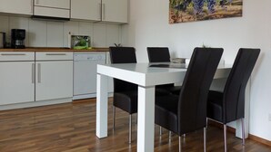 A modern kitchen with white cabinets, a dishwasher, and a coffee maker. There is a dining table with four black chairs and a laptop on it. A painting of grapes hangs on the wall. - HAARDTER WINZER (Whg. 5) renovated and lovingly decorated "Pfalzcard-Partner" (Neustadt an der Weinstraße)