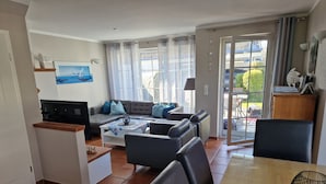 Een moderne woonkamer met een bank, salontafel en eethoek. Er is een televisie, een lamp en uitzicht op het omliggende landschap door grote ramen.