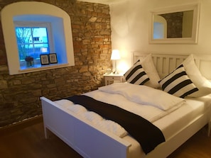 A bedroom with a stone wall, a bed with white bedding and black and white striped pillows, a bedside lamp, and a small table with a clock. - Holiday house situated directly on the water  (Pförring)