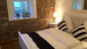 A bedroom with a stone wall, a window with a view, a bedside table with a lamp, and a bed with striped pillows.