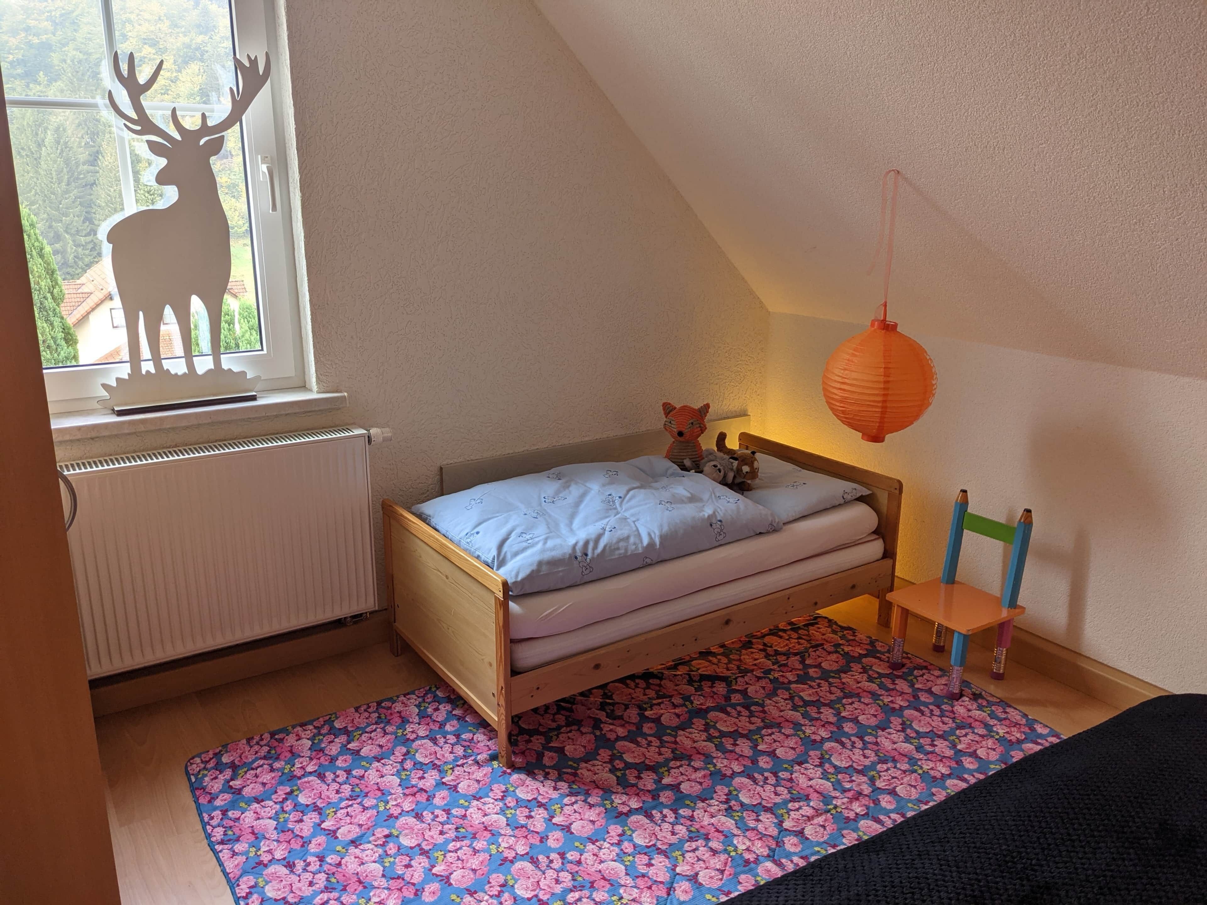 A bedroom with a bed, a window with a deer silhouette, a radiator, a wooden chair, and a red lantern.