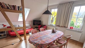 A cozy room with a dining table, chairs, a bookshelf, a red sofa, a TV, and a window with curtains. - Historic villa & comfort in the biosphere reserve (Vesser)