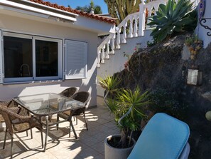 Une terrasse avec une table en verre et des chaises en osier, une plante en pot et un mur blanc agrémenté d’un élément décoratif.