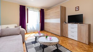 A modern living room with a sofa, two small tables with decorative items, a television on a cabinet, and a large window with curtains. - Two bedrooms Apartment Eva with beautiful garden in Funtana (Funtana)