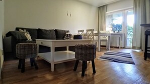 Una sala de estar con un sofá, una mesa blanca, dos taburetes de mimbre, un tapete a rayas y vistas a un balcón con una ventana.
