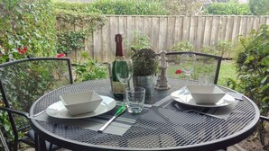 Mesa metálica redonda dispuesta para dos personas, con platos blancos, una botella de vino, copas y una pequeña planta en maceta.
