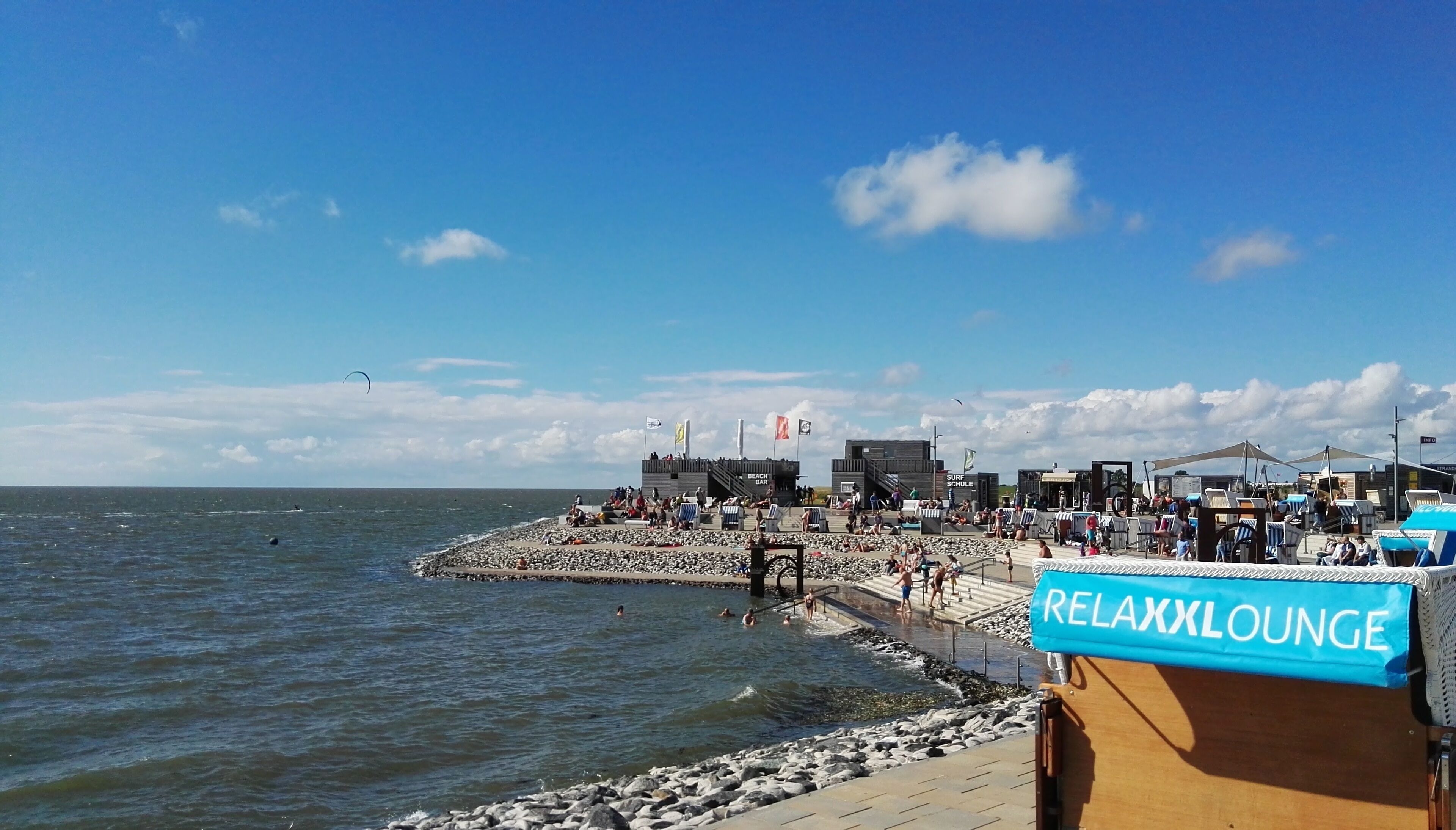Una zona sulla spiaggia con un cartello 'Relaxx Lounge', una costa rocciosa e un molo con persone e bandiere.