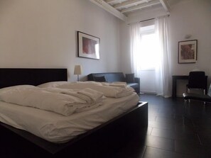 A hotel room with a large bed, a desk, a chair, and two framed pictures on the walls.