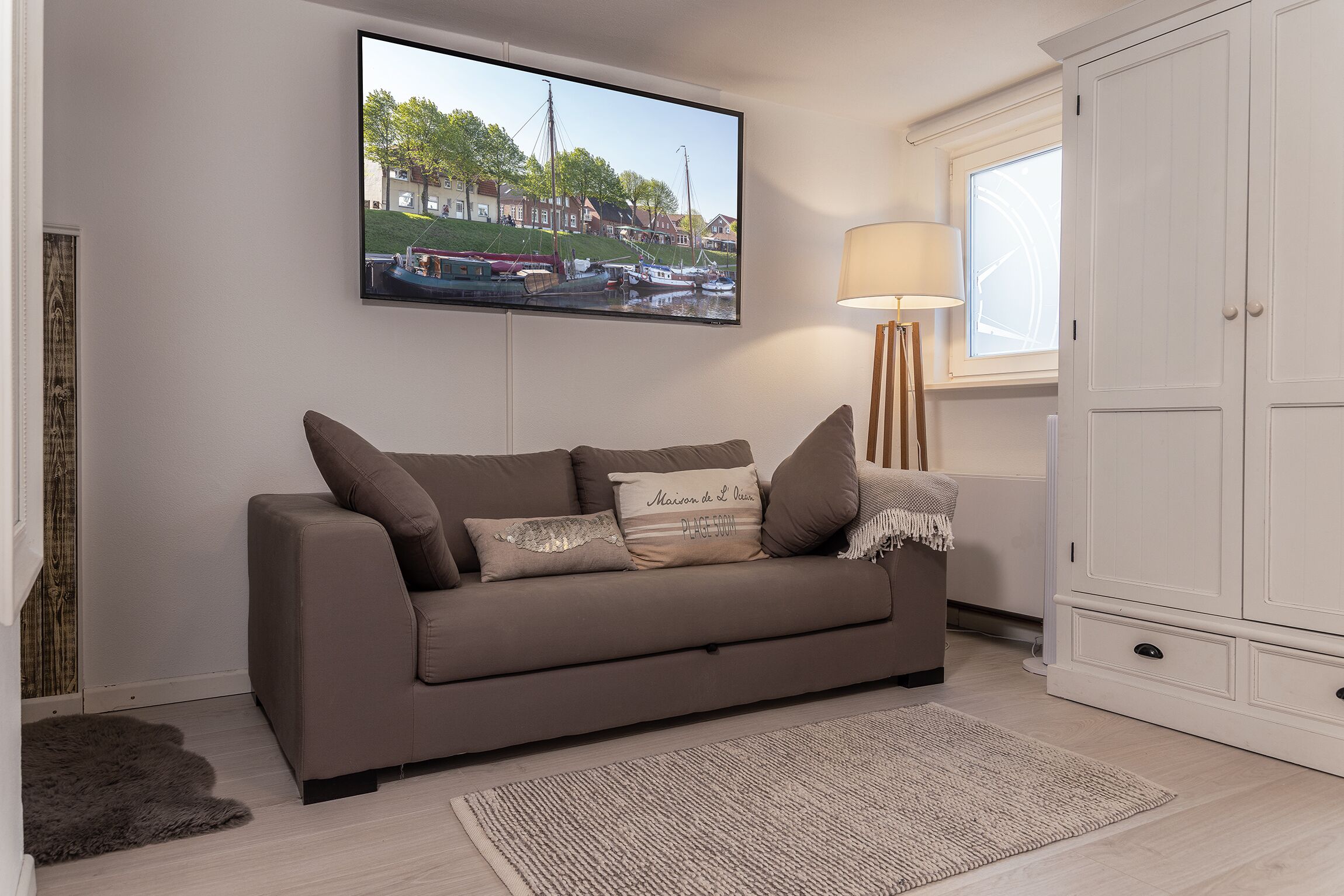 A living room with a brown sofa, a flat-screen TV mounted on the wall, a floor lamp, and a window with a view of houses and boats.