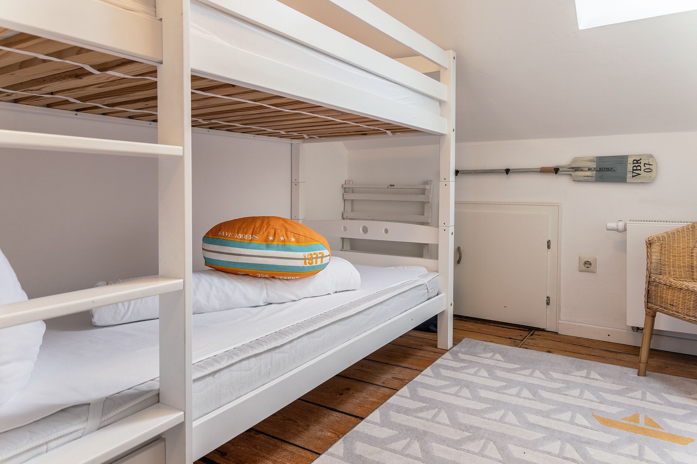 A bunk bed room with a white wall, a wooden floor, and a patterned rug.