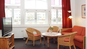Ein gemütliches Zimmer mit Korbmöbeln, einem Fernseher und Blick auf Gebäude durch große Fenster.