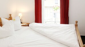 Ein ordentlich bezogenes Bett mit weißen Leinen, ein Nachttisch mit einer Lampe und ein Fenster mit roten Vorhängen.