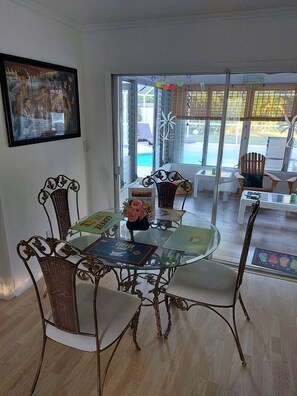 A dining area with a glass table surrounded by wicker chairs, a framed picture on the wall, and a view of a pool through large windows. - Top location, heated pool, Wi-Fi, Florida Room, Tropical Garden (Cape Coral)