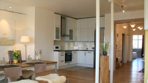 Une cuisine moderne avec des meubles blancs, un coin repas avec une table et des bancs en bois, et un couloir menant à une chambre.
