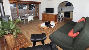 Sala de estar com sofá, mesa de centro, televisão e lareira. Há também uma área de jantar com mesa e cadeiras, e uma cozinha visível por um arco.