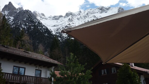Vista de montanhas com picos nevados, um chalé de madeira com varanda e um guarda-sol grande.