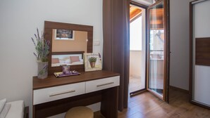 Een slaapkamer met een houten bureau, een bed en een klein krukje. Er is een raam met gordijnen, een deur en uitzicht op een balkon.