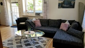Un salón con un sofá seccional gris oscuro, una mesa de centro de vidrio y una alfombra estampada. Hay una ventana con cortinas, una silla y un cuadro en la pared.