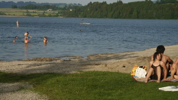 Escena junto al lago con gente nadando y descansando en la orilla.