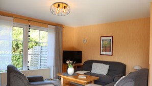 Un salon comprenant un canapé, deux fauteuils, une table basse, un téléviseur et une fenêtre avec des rideaux.