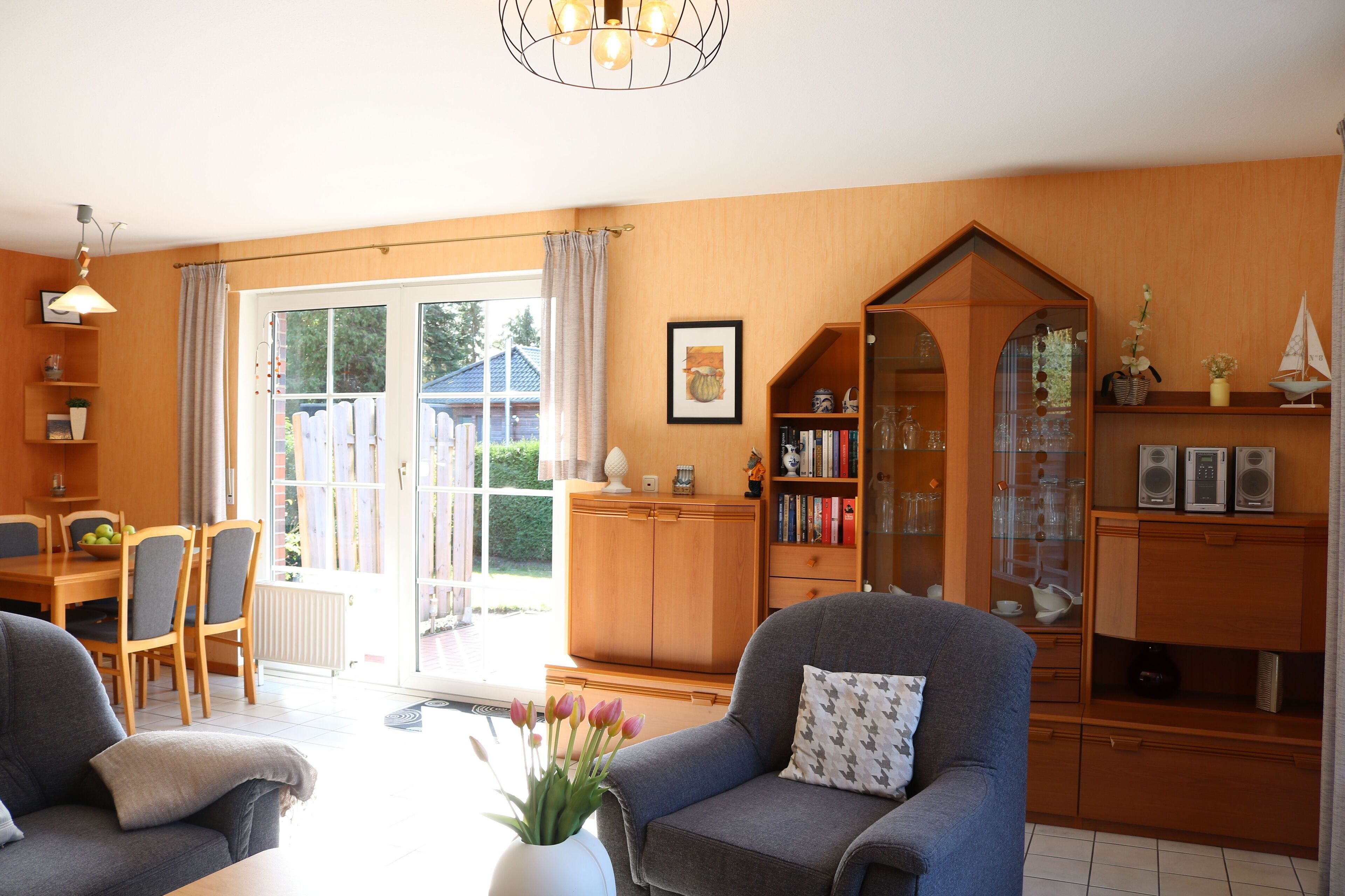 A cozy living room with a sofa, armchair, and a dining area with a table and chairs. There is a bookshelf and a cabinet with glass doors.