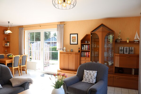 A cozy living room with a sofa, armchair, and a dining area with a table and chairs. There is a bookshelf and a cabinet with glass doors.