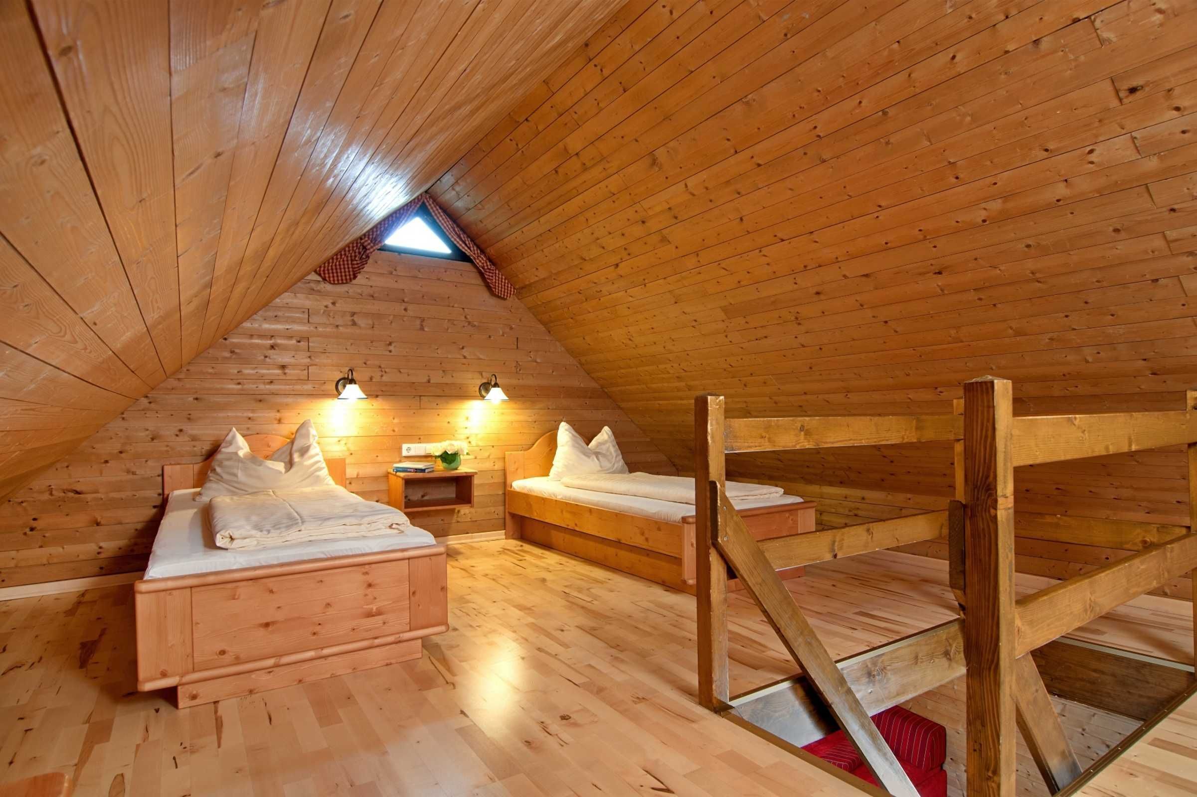 Een knus zolderkamer met houten wanden en vloeren, twee bedden, een kleine tafel en een raam met een rood geruit gordijn.