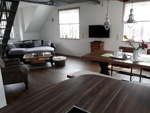 Un salon moderne avec un canapé, une table basse et une table à manger. Un téléviseur est fixé au mur, il y a un meuble en bois et une fenêtre avec des stores.