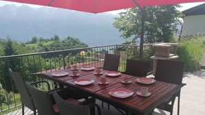 A dining table set for six with wicker chairs, a red umbrella, and a scenic mountain view.